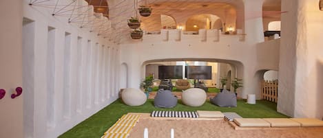 Een ruime woonkamer met een hoge plafond, hangplanten en een gezellige zithoek met kussens en een loungestoel.