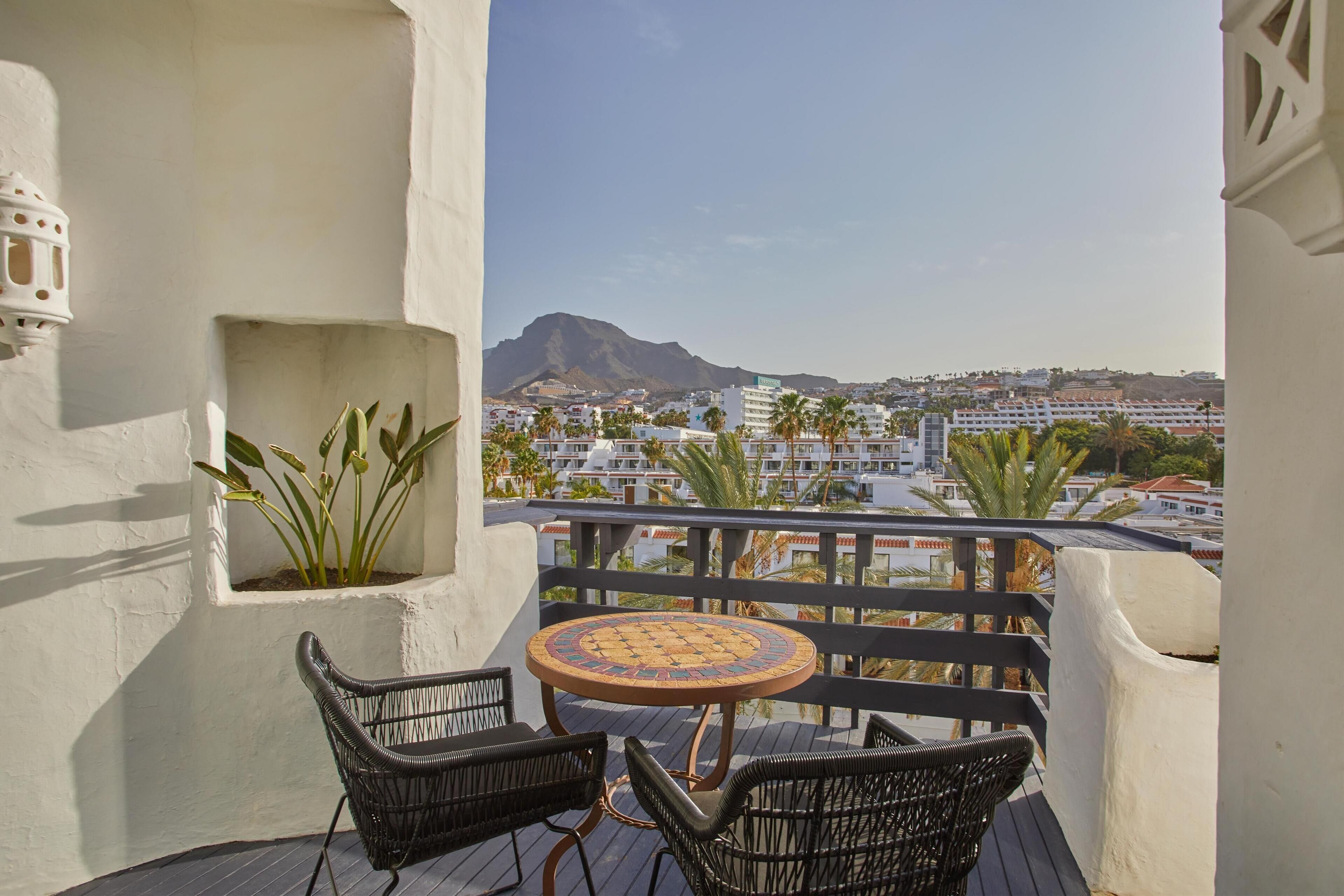 Een balkon met rieten stoelen en een ronde tafel, met uitzicht op een stadsbeeld met gebouwen en een berg.