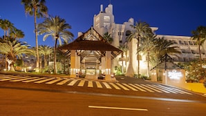 Entrada de hotel con un corredor cubierto, palmeras y un edificio distintivo al fondo.