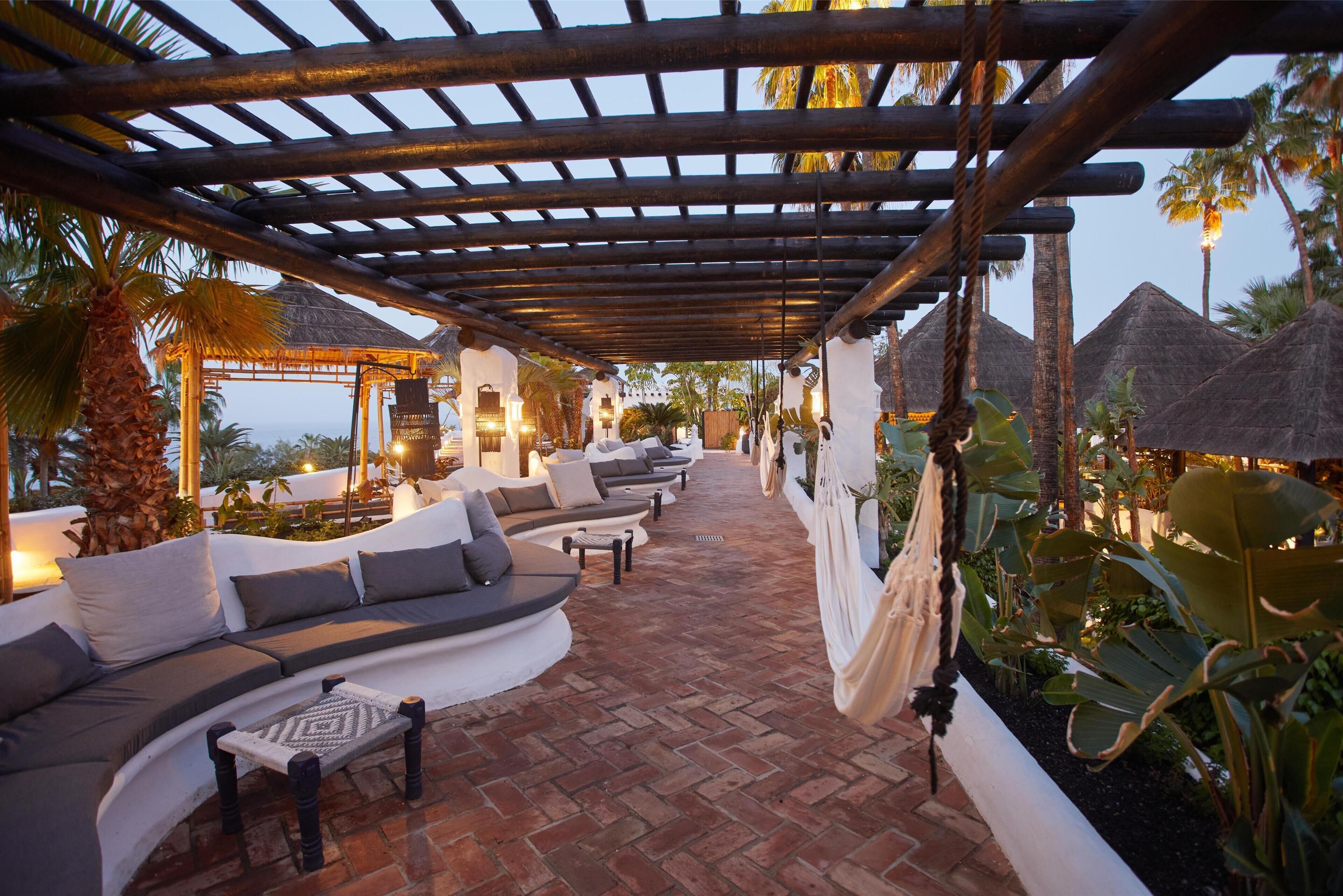 A covered outdoor seating area with cushioned benches and a patterned rug, surrounded by tropical plants and palm trees.