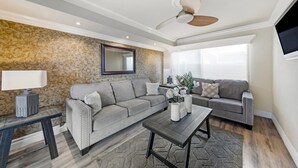 A living room with a grey sectional sofa, a coffee table, a TV mounted on the wall, and a ceiling fan.