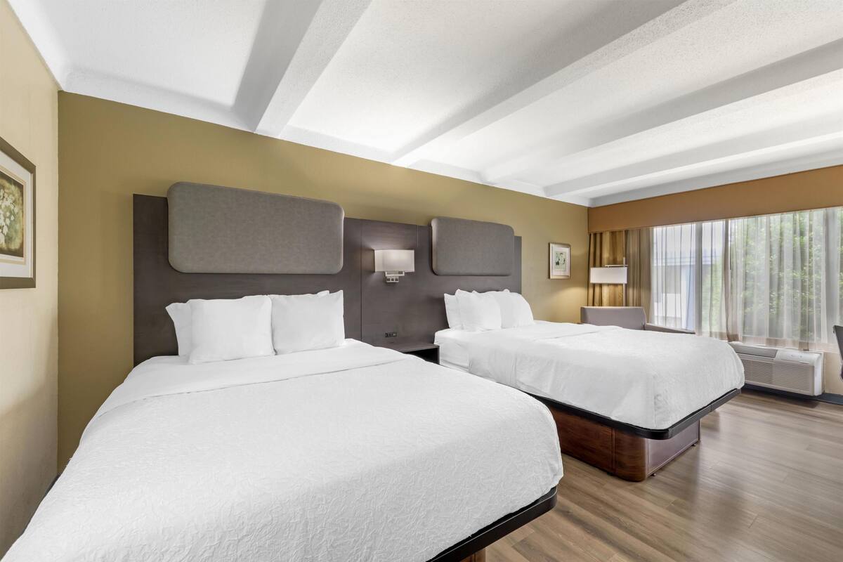 Une chambre d’hôtel avec deux lits, une grande fenêtre avec des rideaux, un plafond doté de lucarnes et une lampe fixée au mur.