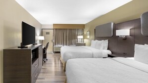 Habitación de hotel con dos camas, un escritorio con televisor, una lámpara y una luz empotrada en la pared.