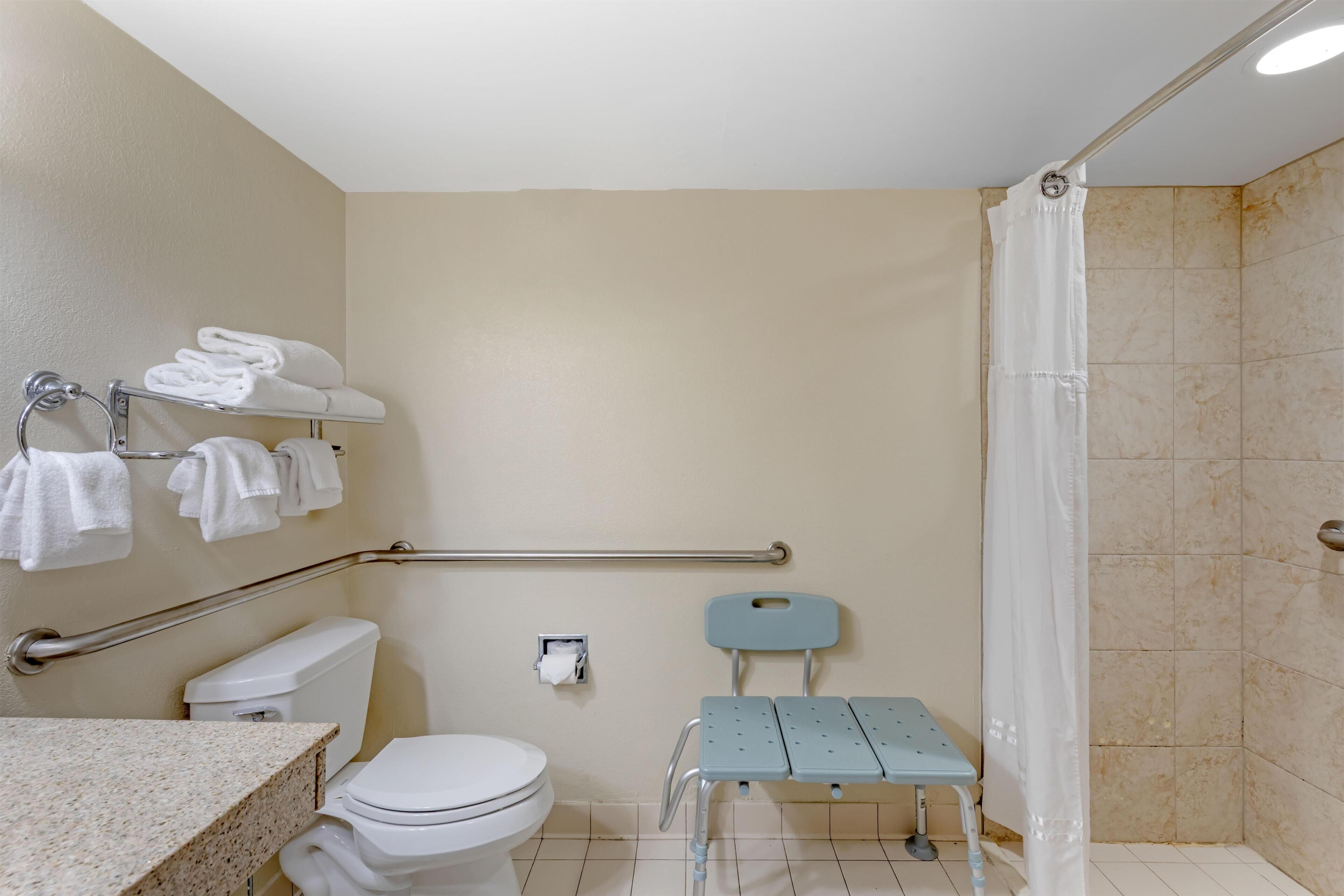 A bathroom with a toilet, a sink, a shower, a grab bar, a chair, and towels on a rack.