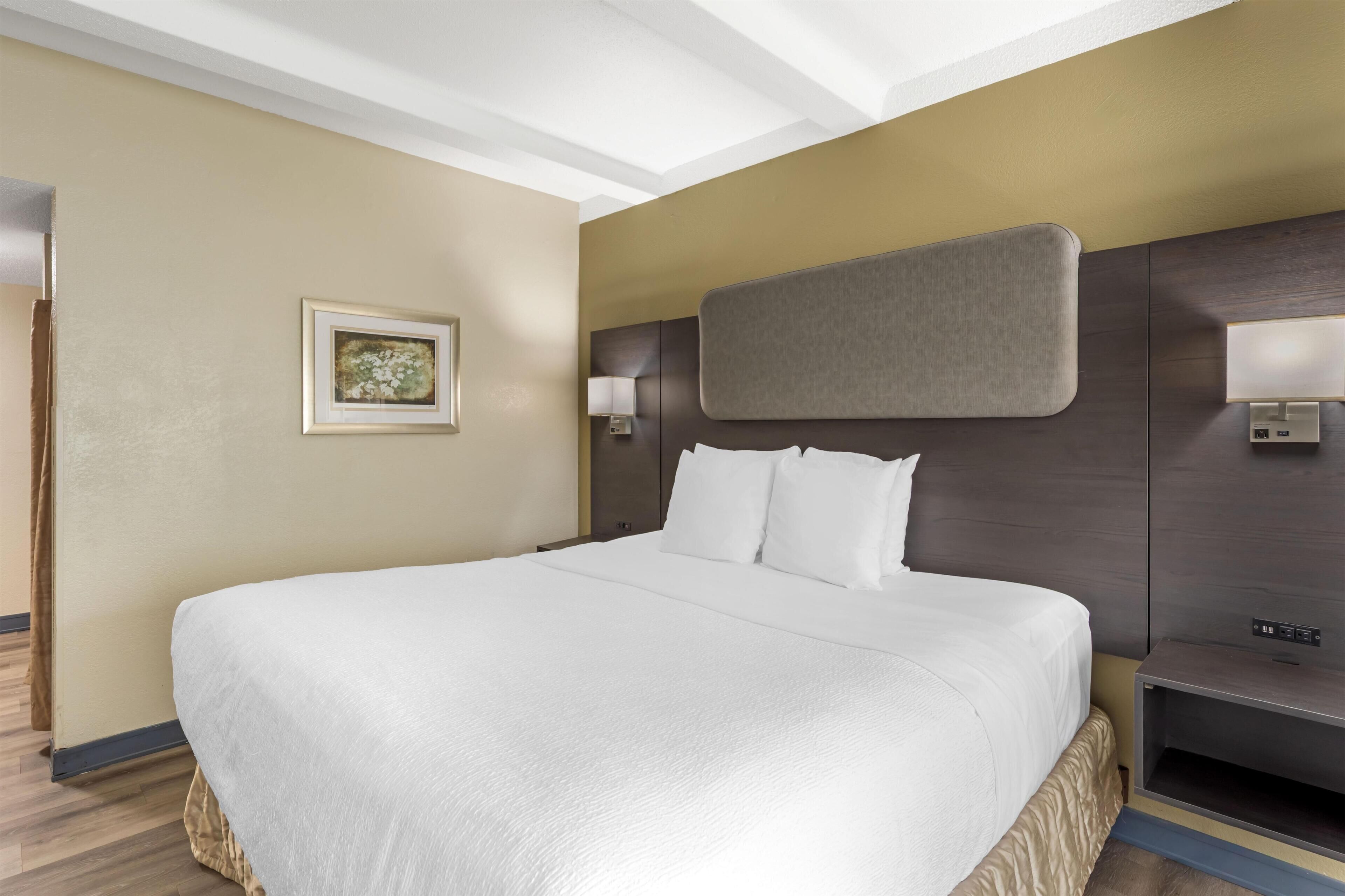 A hotel room with a large bed, a headboard with a control panel, a bedside table, and a wall-mounted lamp.