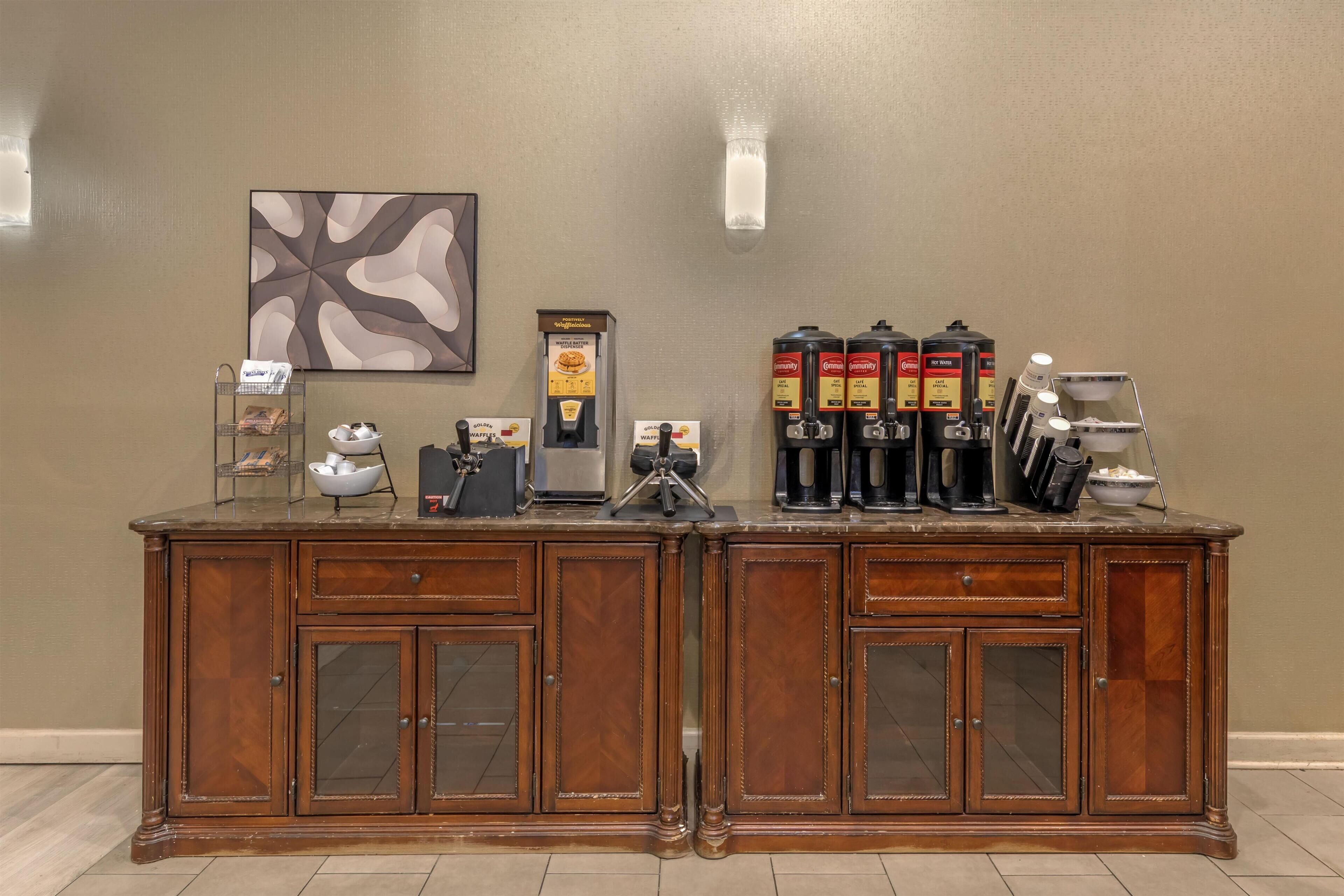 A wooden buffet with a coffee station, including a waffle maker, coffee makers, and condiment dispensers.