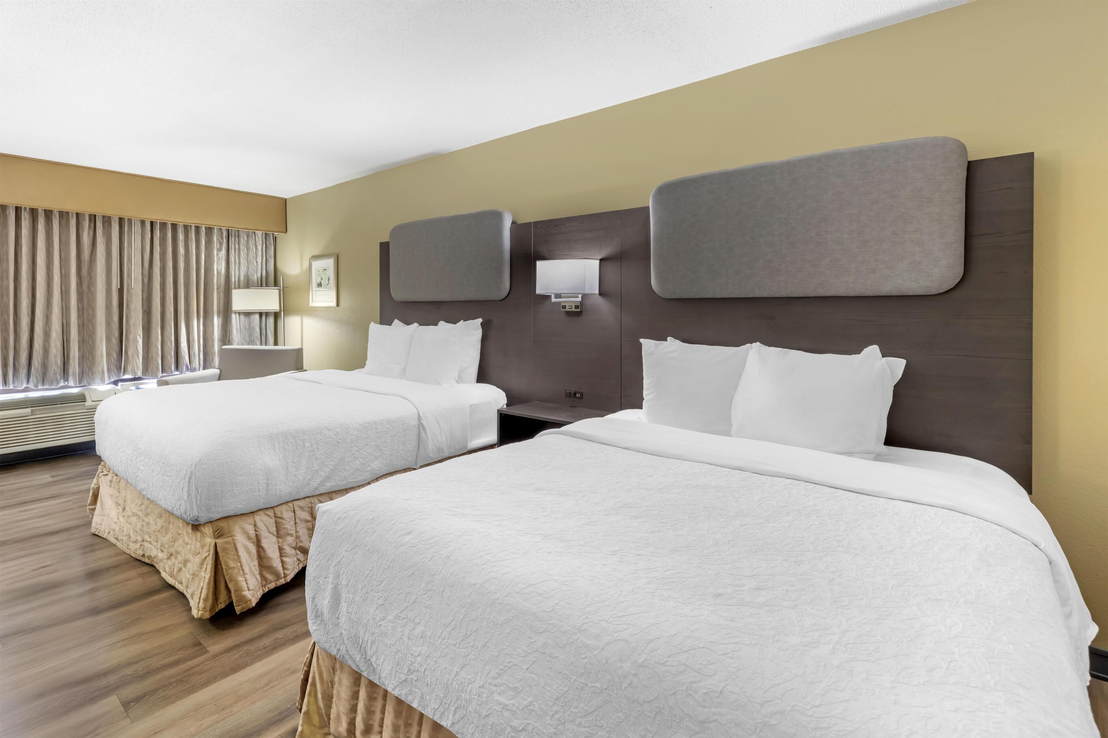A hotel room with two beds, a large window with curtains, a wall-mounted lamp, and a wooden floor.