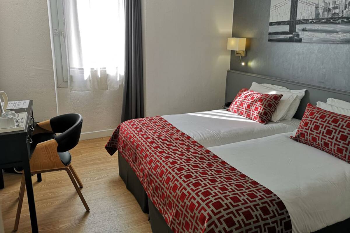 Een hotelkamer met twee bedden, een bureau met stoel, een raam met gordijnen en een wandlamp.