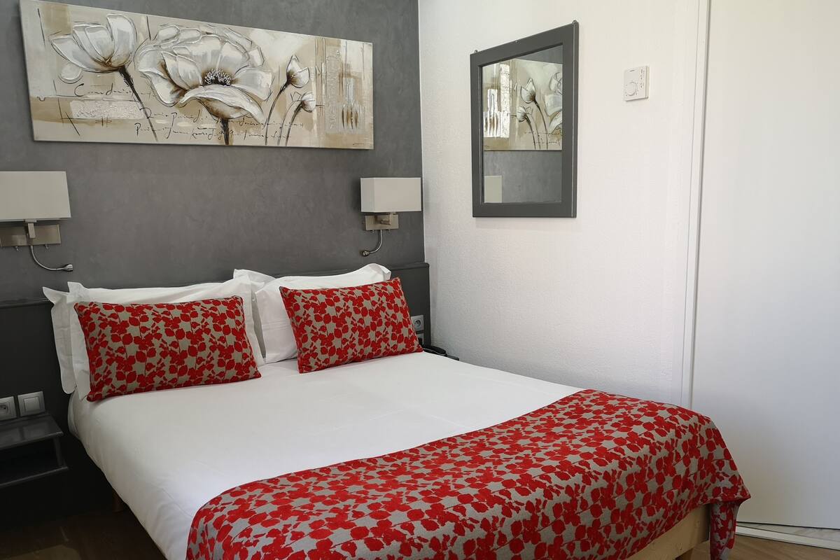 Een bed met witte beddengoed en rode kussens met bloemenprint, een schilderij erboven en een spiegel aan de muur.