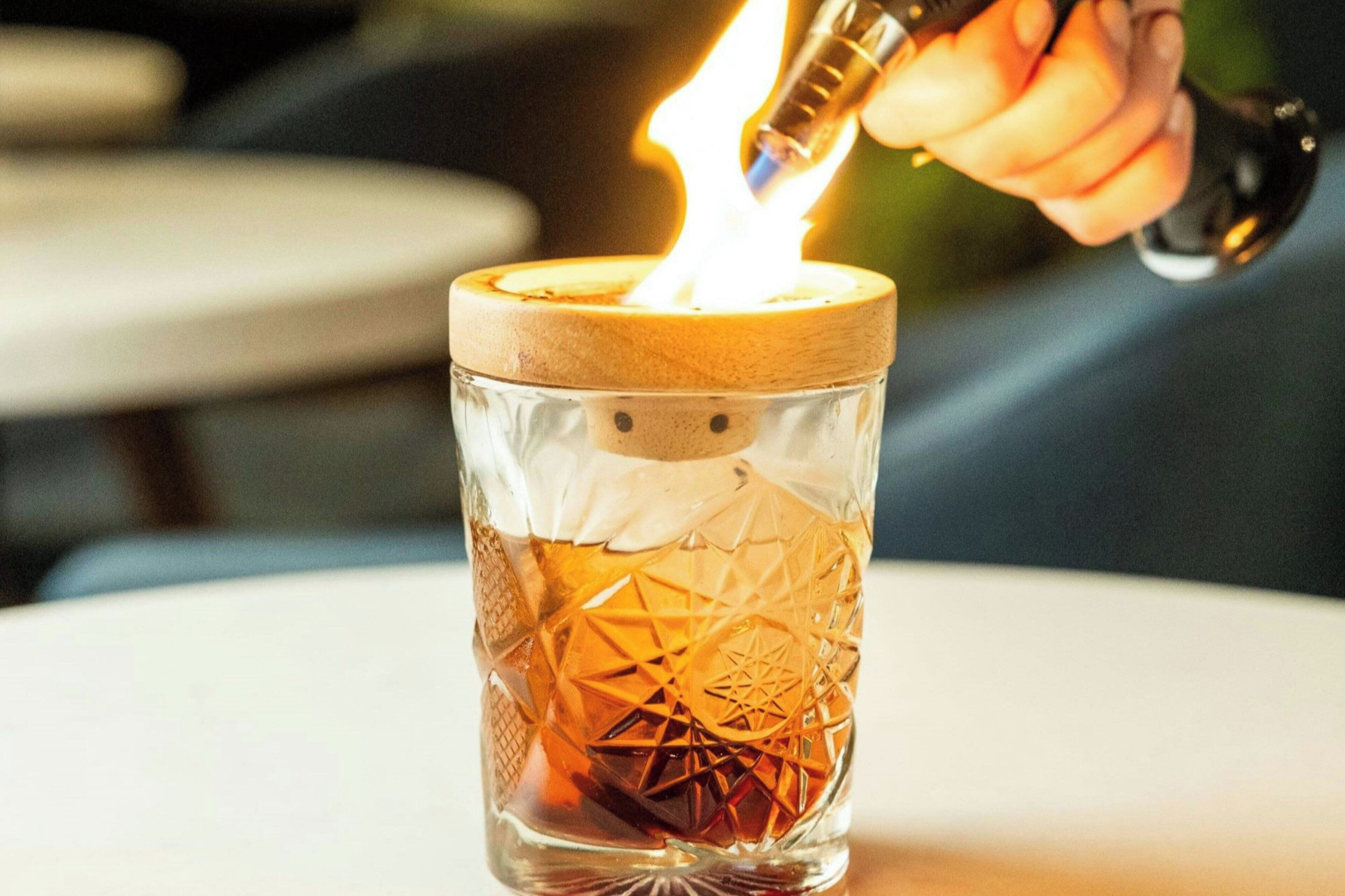 Une bougie dans un support en verre, recouverte d’un couvercle en bois, que quelqu’un est en train d’allumer.