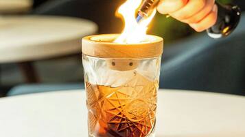 Une bougie dans un support en verre, recouverte d’un couvercle en bois, que quelqu’un est en train d’allumer.