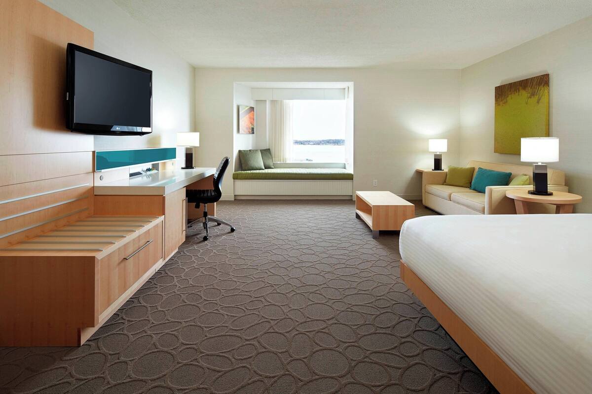 A hotel room with a bed, a desk with a chair, a TV mounted on the wall, a window with a view, and two bedside tables with lamps.