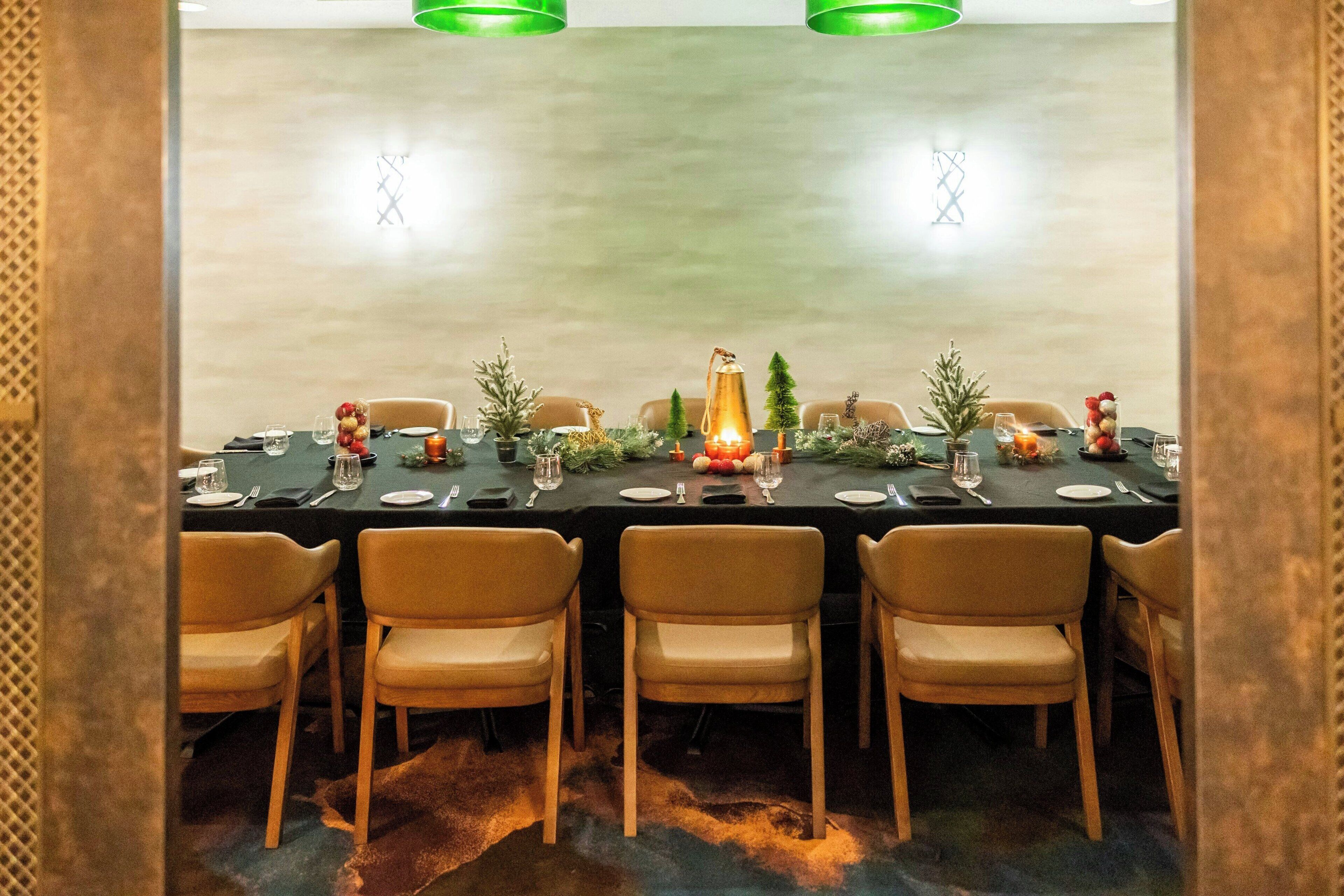 A dining area with a long table set for six, surrounded by six chairs. The table is decorated with a centerpiece, candles, and small plants.