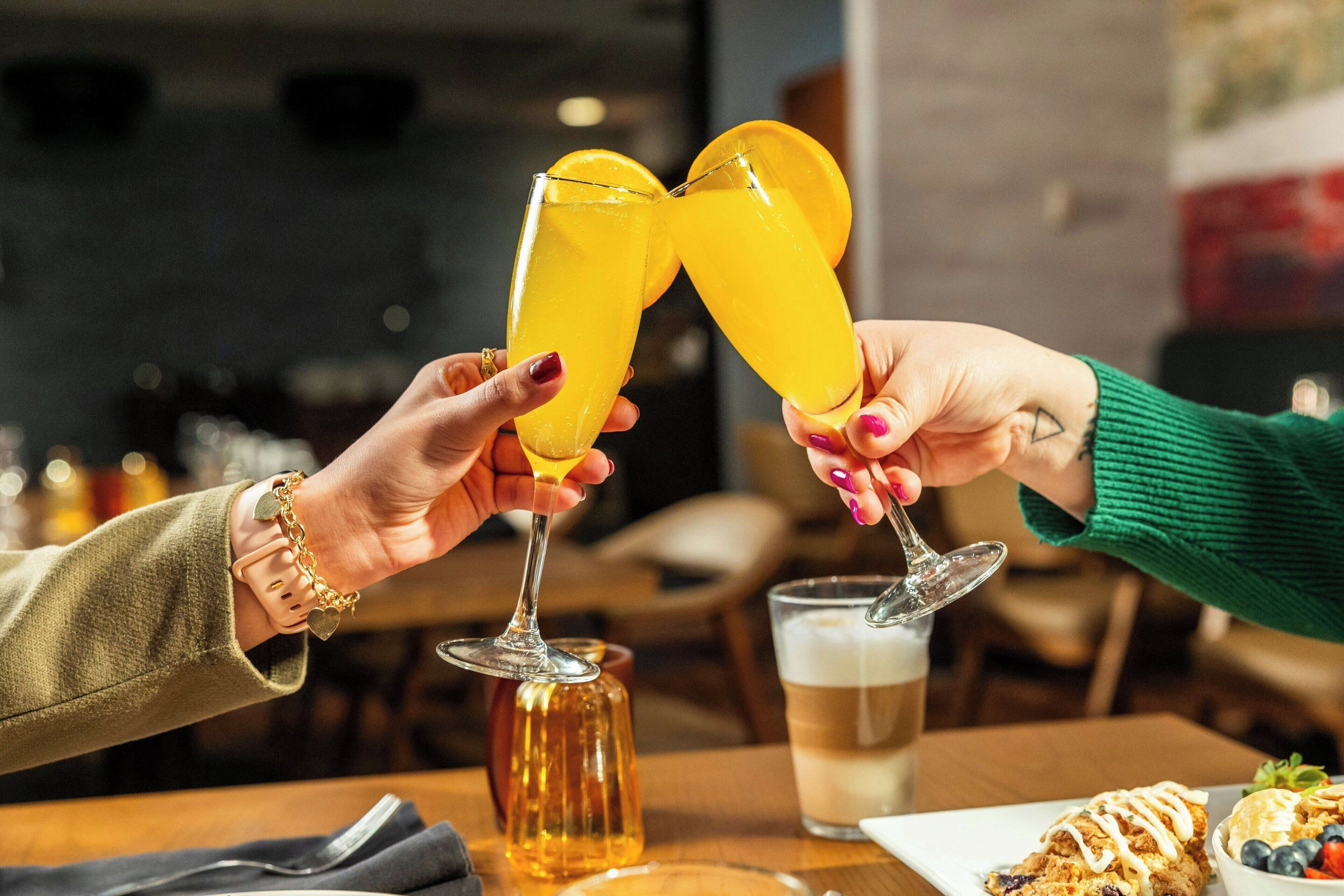 Dos personas brindando con bebidas amarillas, un vaso de cerveza y un plato de comida sobre una mesa.