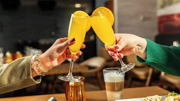 Dos personas brindando con bebidas amarillas, un vaso de cerveza y un plato de comida sobre una mesa.