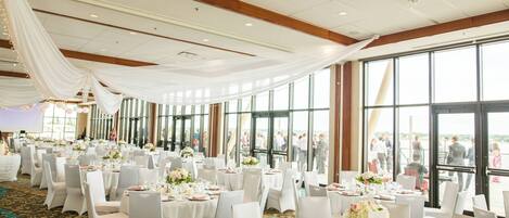 A banquet hall with round tables set for a formal event, large windows, and a high ceiling with draped fabric.