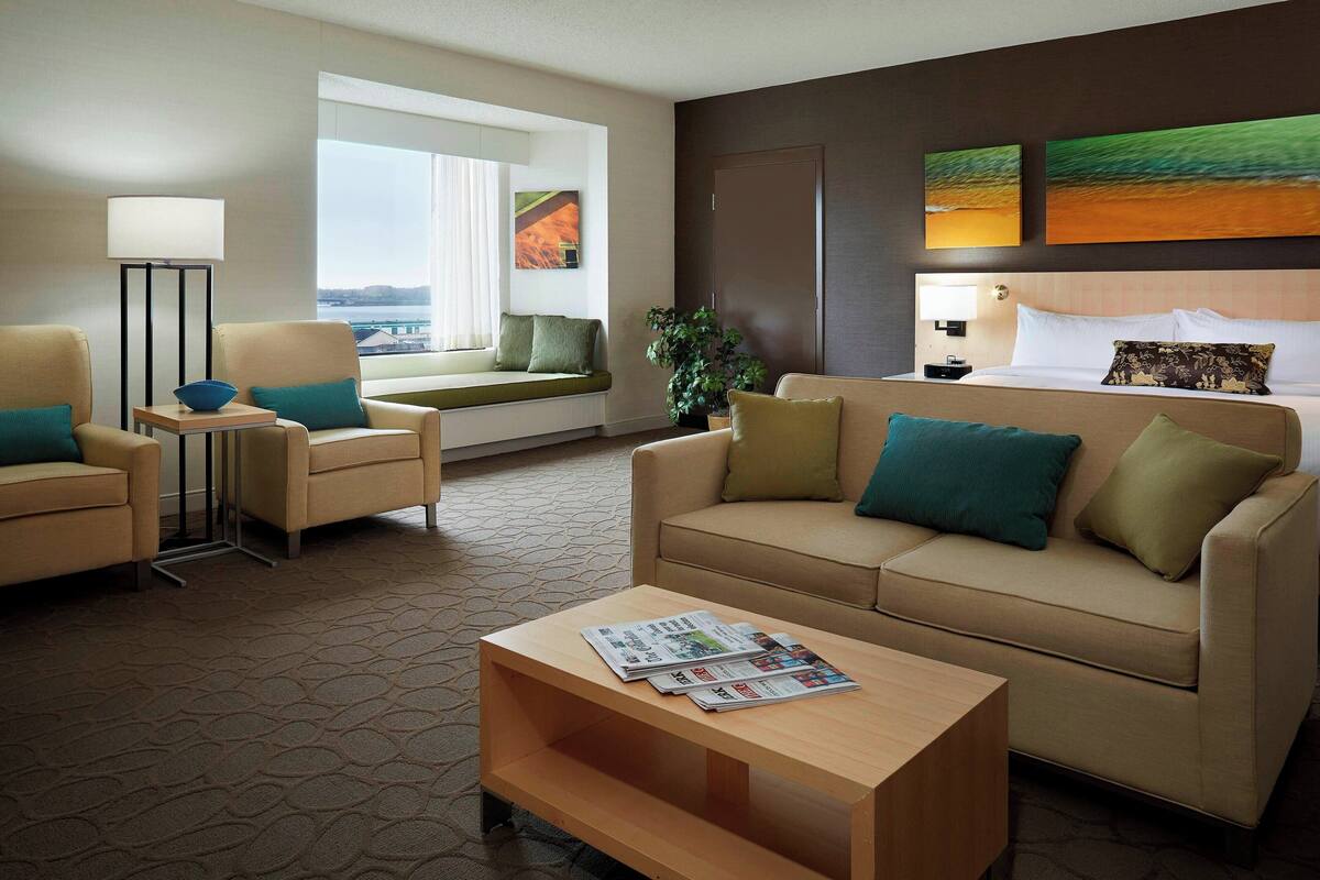 A hotel room with a sofa, armchairs, a coffee table with newspapers, a floor lamp, a window with a view, and two paintings on the wall.