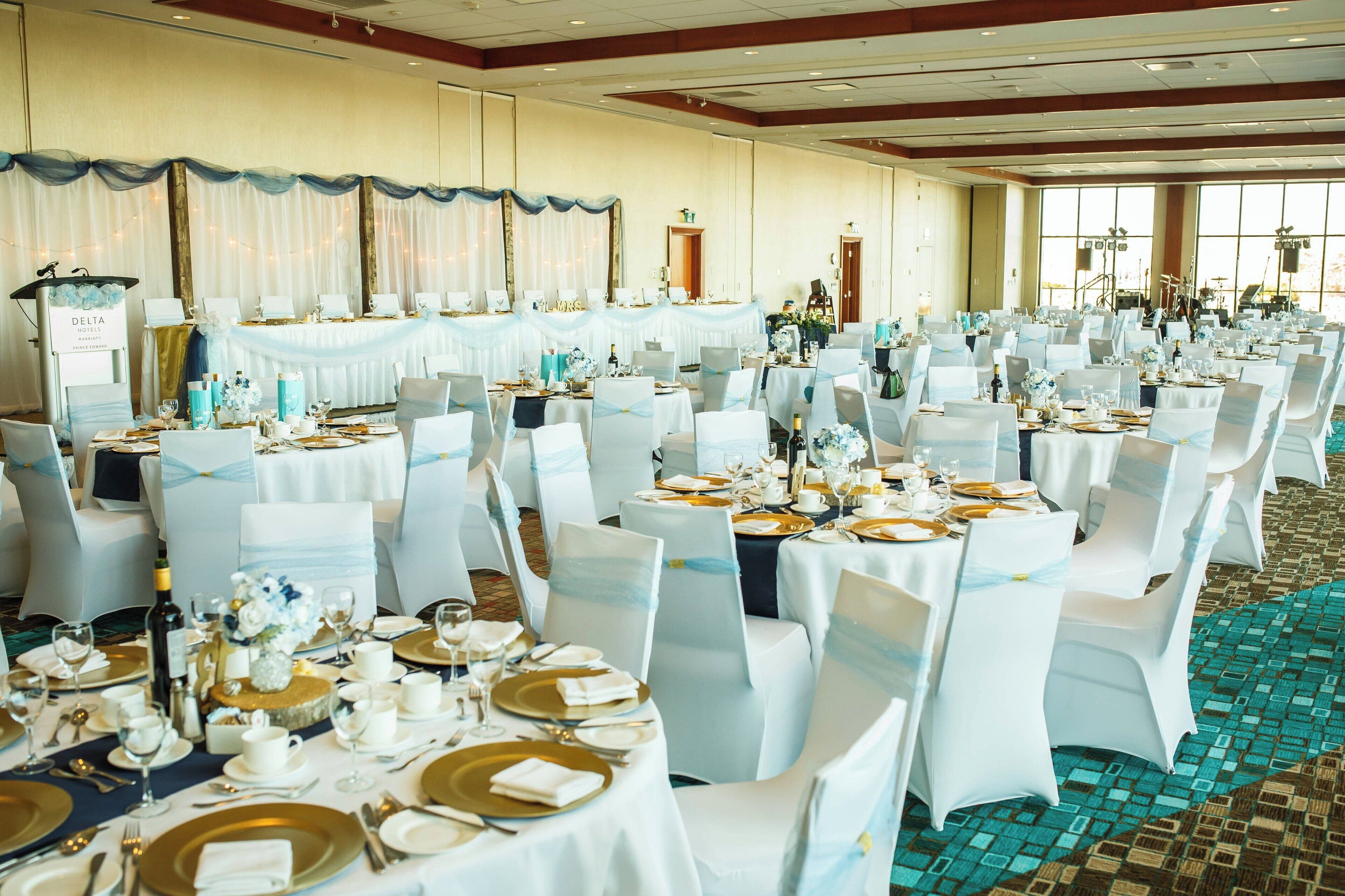 Un salón de banquetes con mesas redondas dispuestas para un evento formal, con manteles blancos, platos bajos dorados y detalles en azul.
