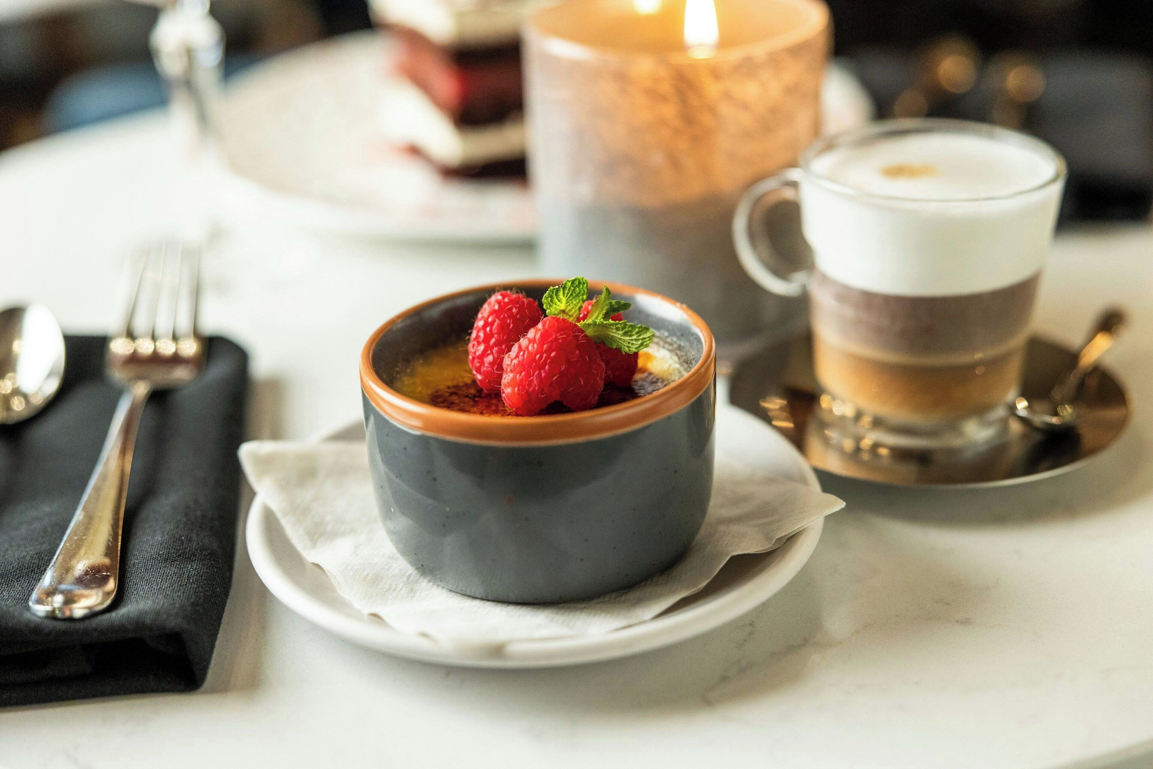 Un tazón de postre con frambuesas y menta sobre un plato blanco, una vela y una taza de café en una mesa.