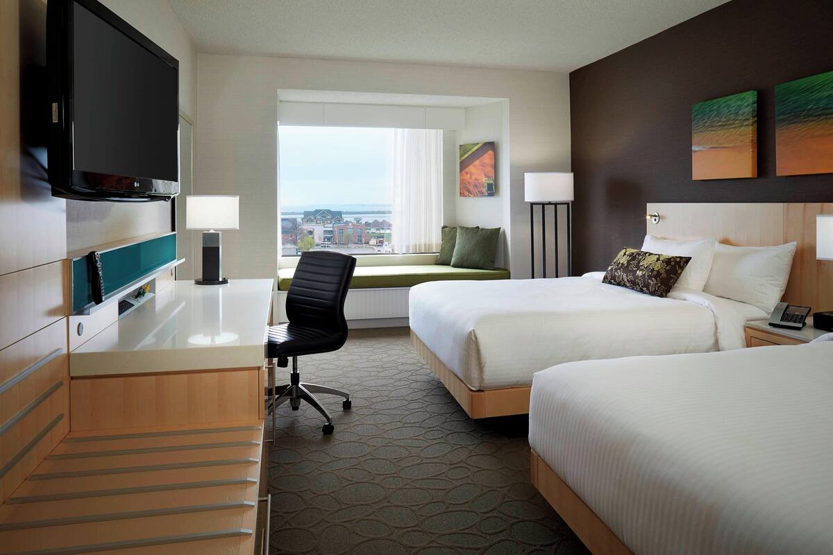 A hotel room with two beds, a desk with a flat-screen TV, a chair, a window with a view of buildings, and artwork on the wall.