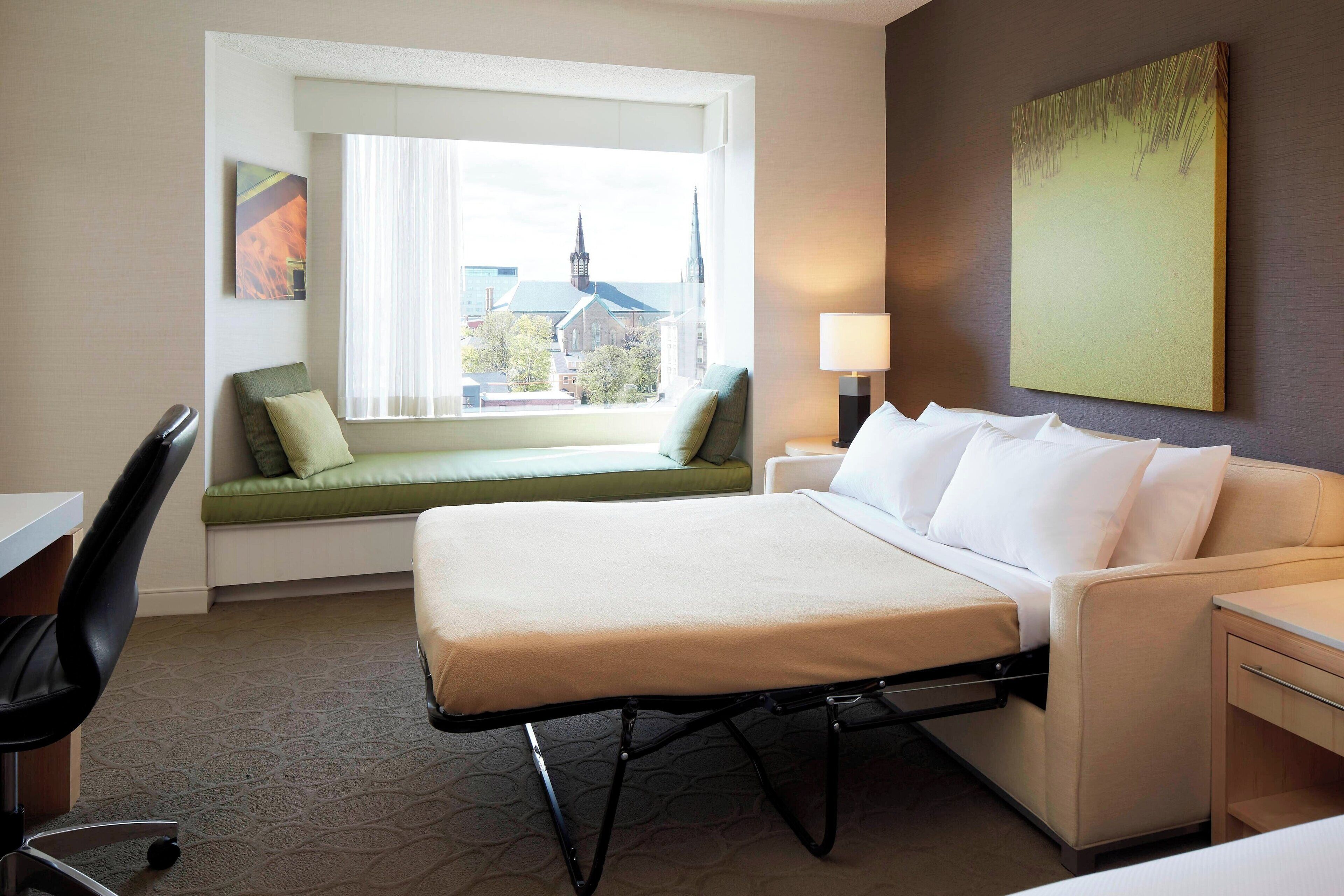 Una habitación de hotel con cama, escritorio, silla, asiento junto a la ventana y lámpara.
