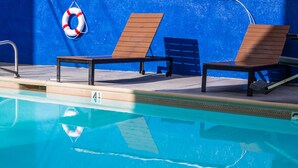 A poolside area with two wooden lounge chairs, a lifebuoy, and a pool with clear blue water. - Anaheim Carriage Inn (Anaheim)