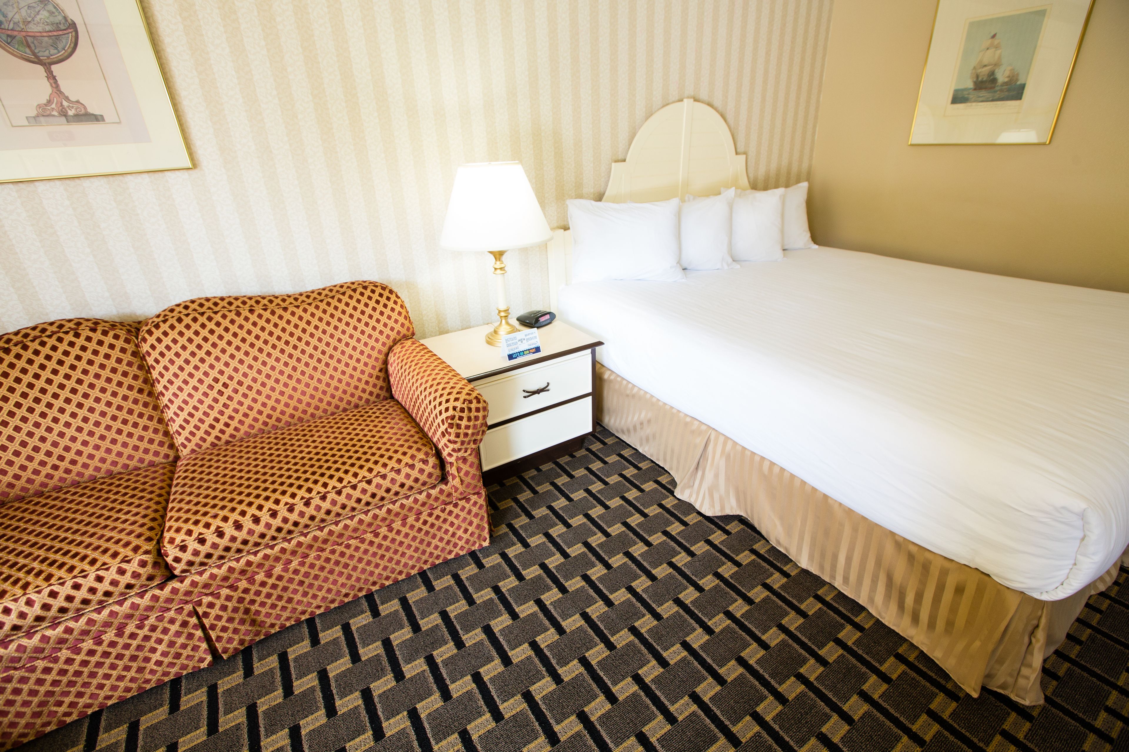 Quarto de hotel com sofá estampado, cama com lençóis brancos, mesa de cabeceira com abajur e quadro na parede.