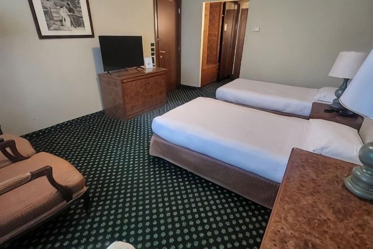 A hotel room with two beds, a television on a wooden stand, a desk with a lamp, and a patterned carpet.