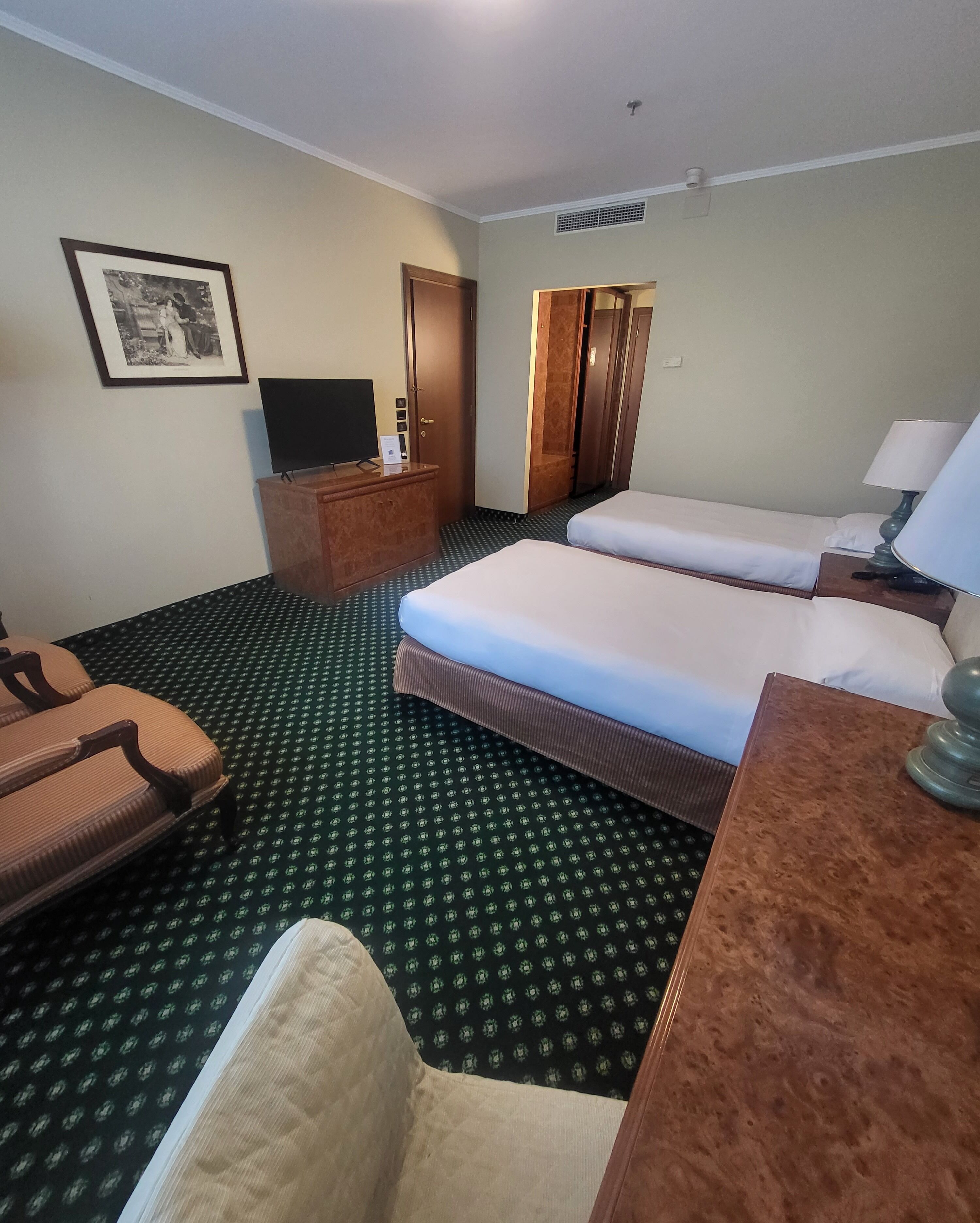 Een hotelkamer met twee bedden, een bureau, een stoel, een televisie en een ingelijst schilderij aan de muur.