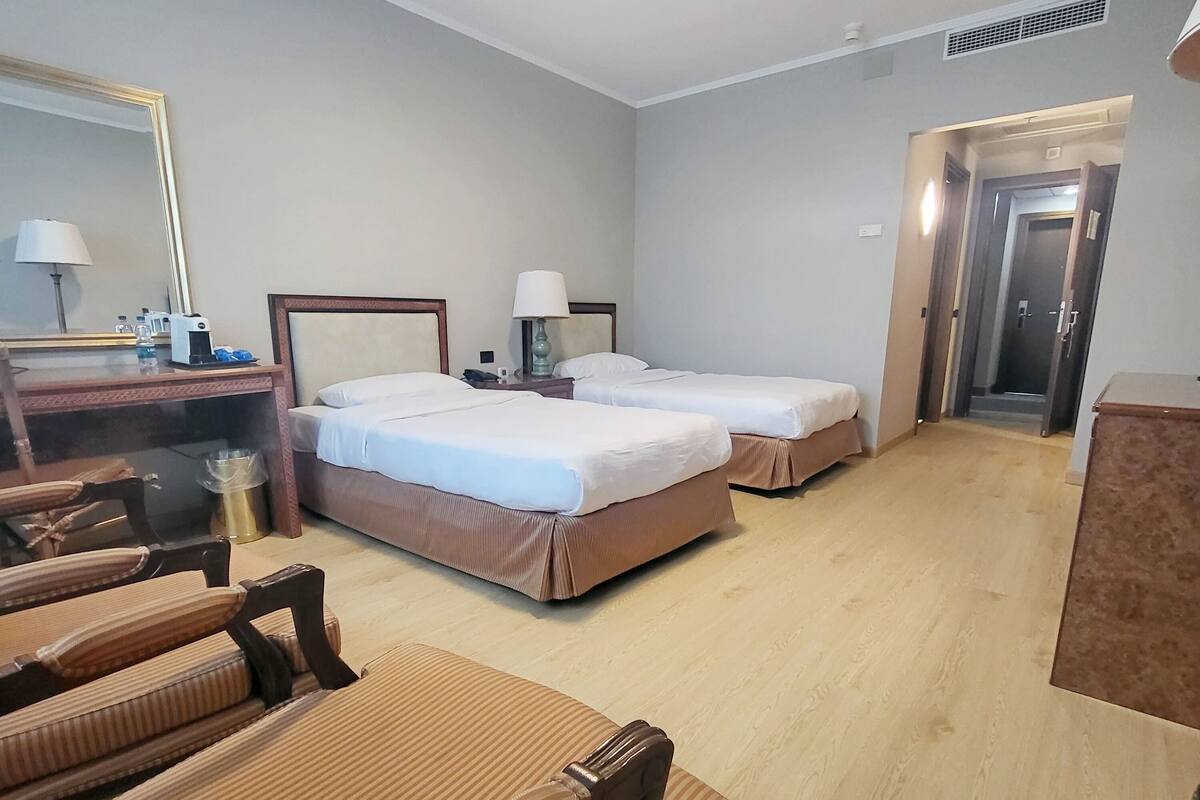 A hotel room with two single beds, a desk with a mirror, a chair, and a small table with a lamp.
