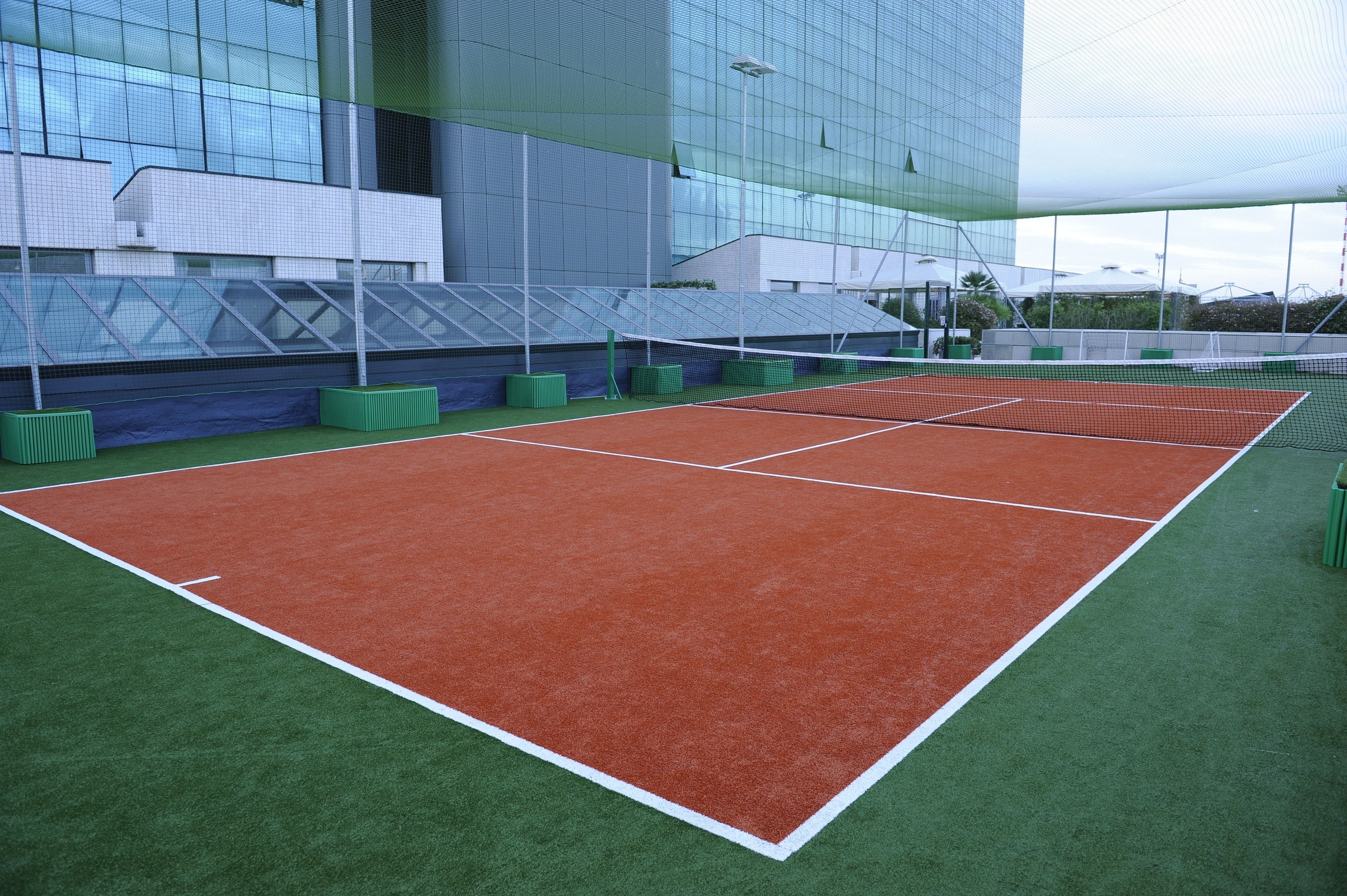 Una cancha de tenis con superficies verde y roja, rodeada por un edificio con grandes ventanales.