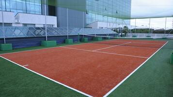 Una cancha de tenis con superficies verde y roja, rodeada por un edificio con grandes ventanales.