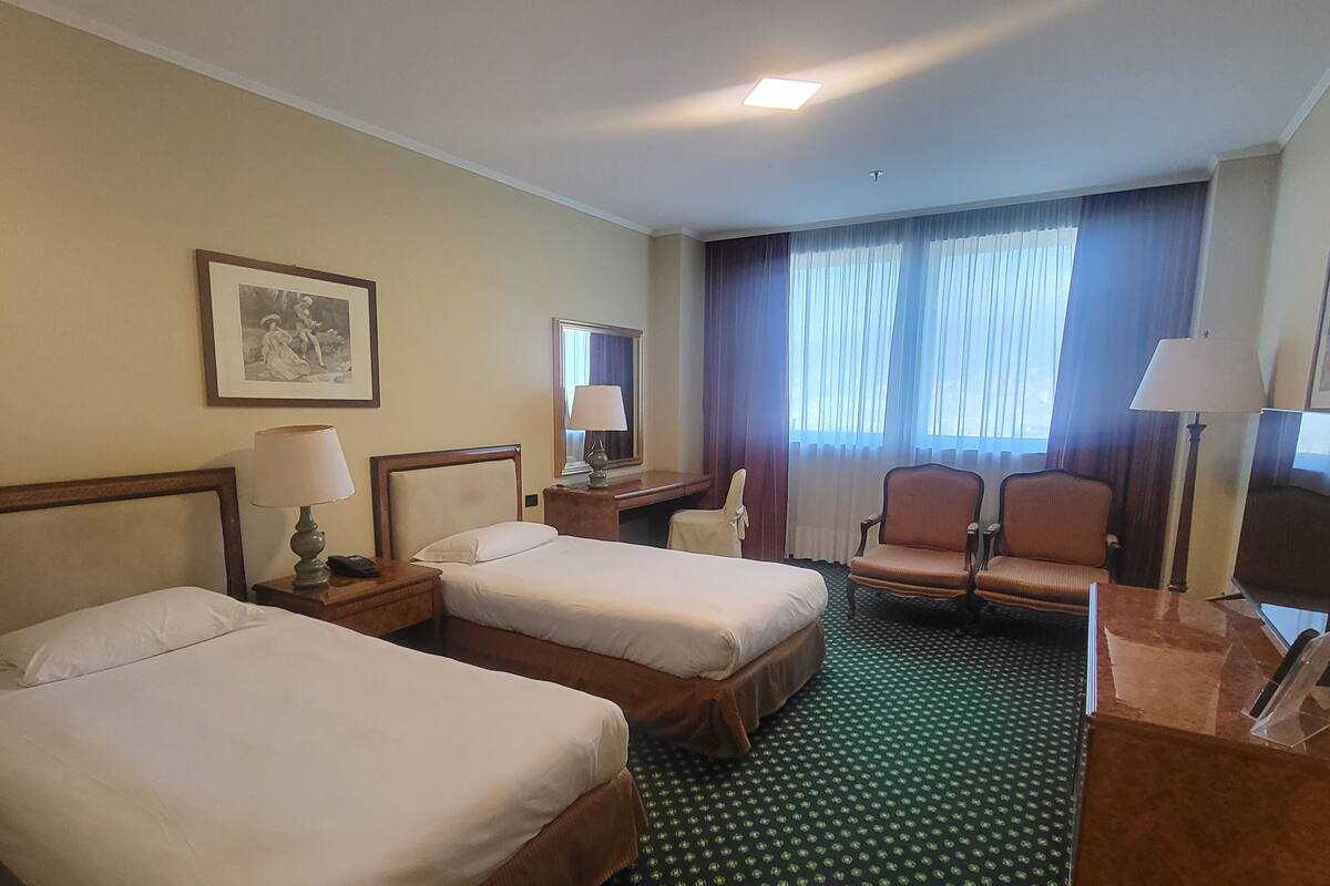 A hotel room with two beds, a desk, a chair, a lamp, a framed picture on the wall, and a window with curtains.