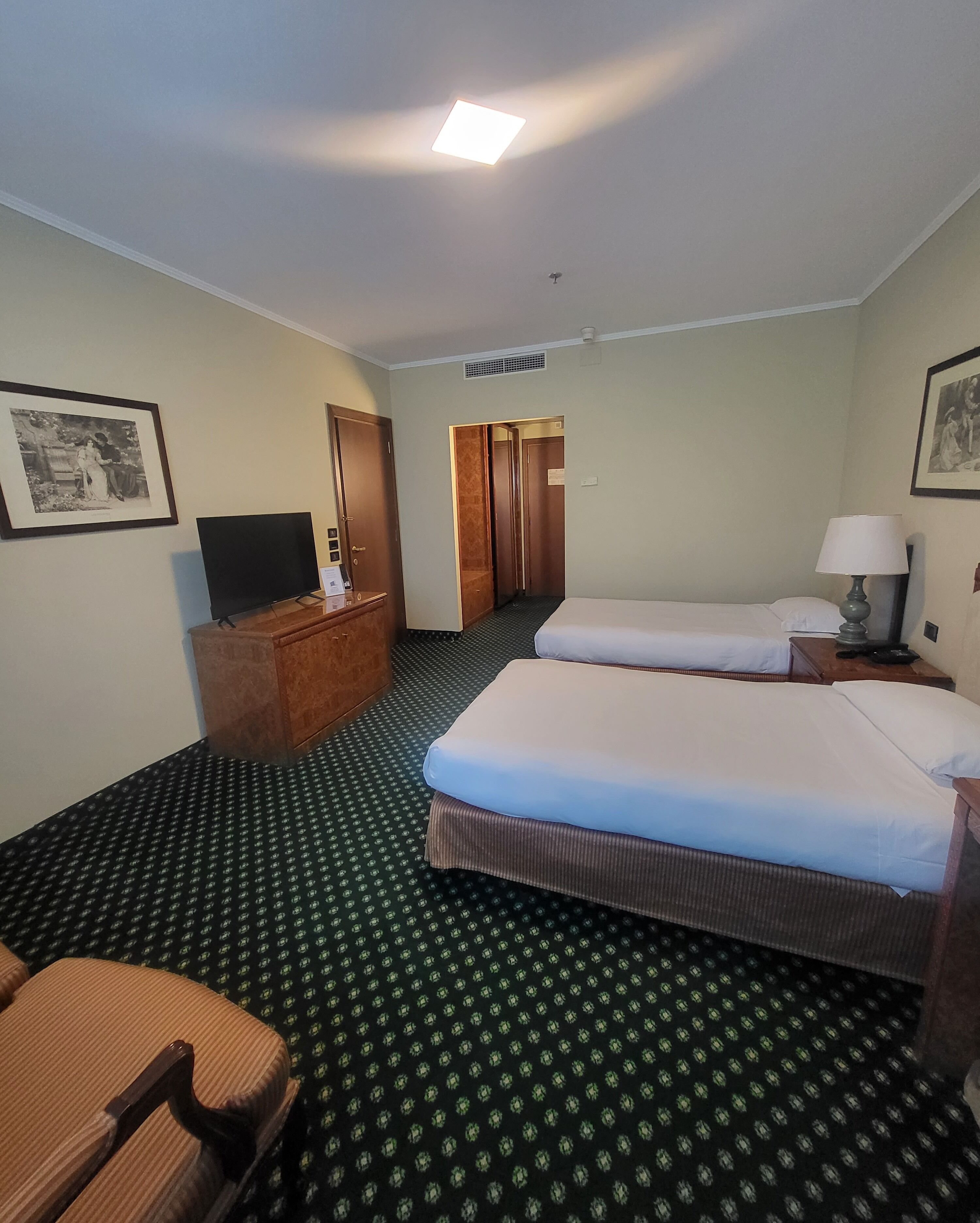 A hotel room with two beds, a desk, a chair, a television, and framed pictures on the walls.