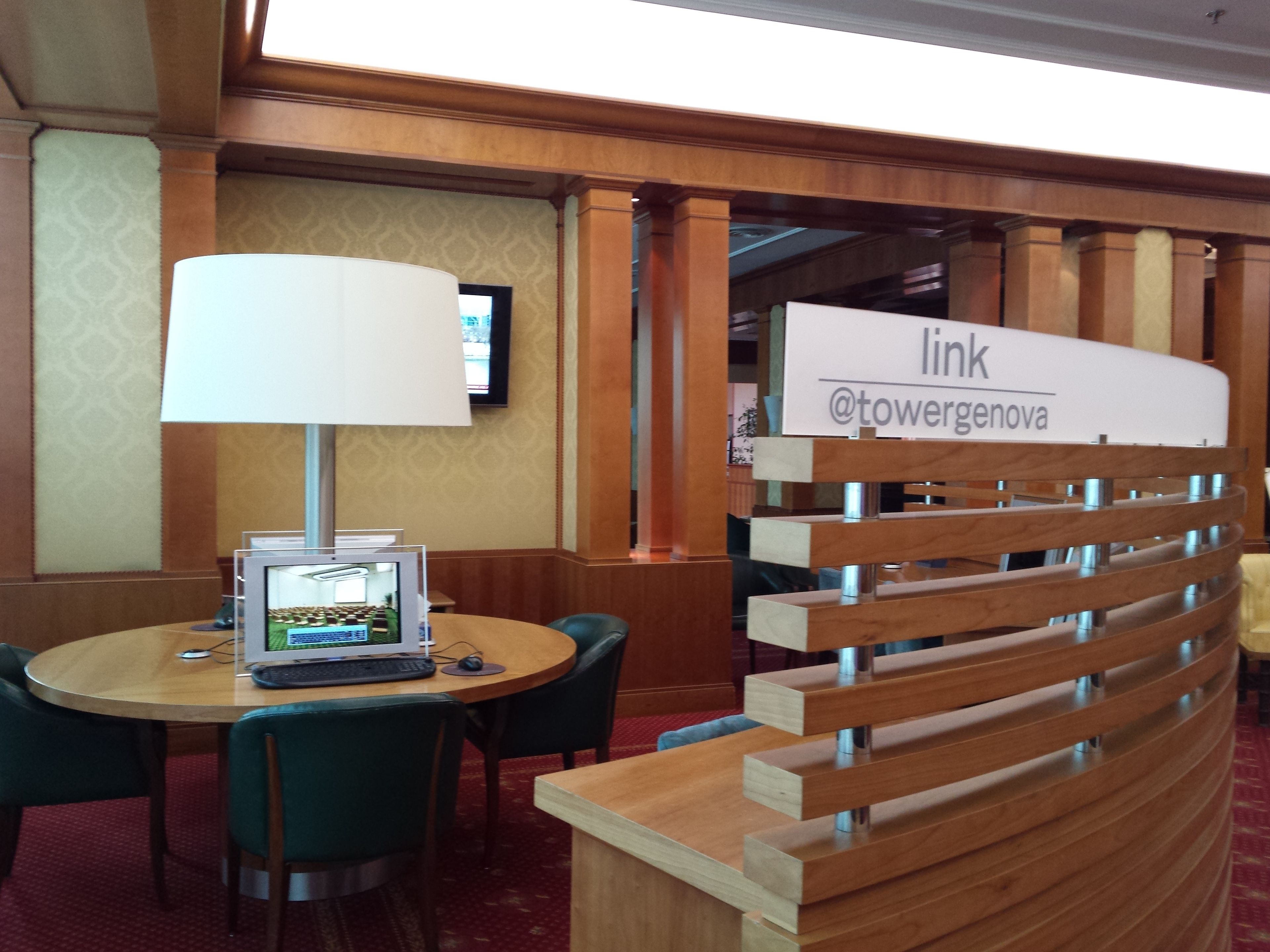 A hotel reception area with a desk, chairs, a lamp, and a sign with the hotel's name and social media handle.