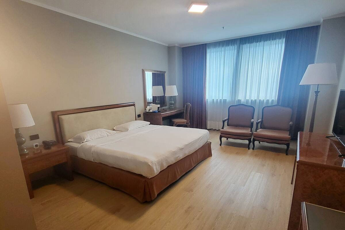 A hotel room with a large bed, a desk, two chairs, a mirror, and a TV.