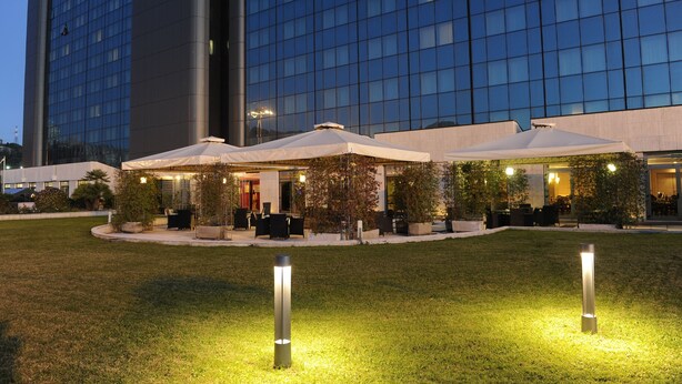 A hotel with a modern glass facade, outdoor seating with umbrellas, and a well-maintained lawn. - Tower Genova Airport Hotel & Conference Center (Genoa)