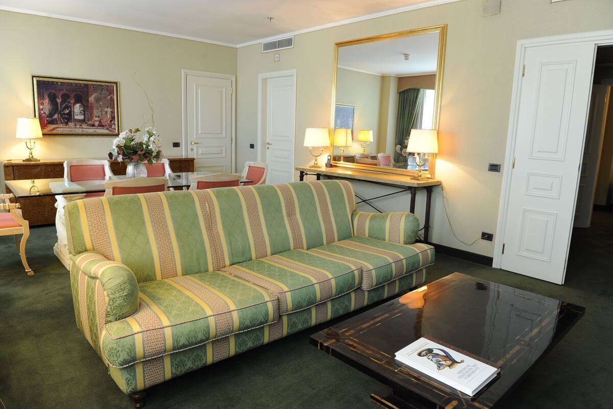A hotel room with a striped sofa, a glass coffee table, a desk with lamps, a painting on the wall, and a large mirror.