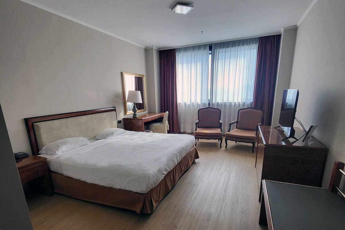 A hotel room with a large bed, a desk, two chairs, a TV, and a window with curtains.
