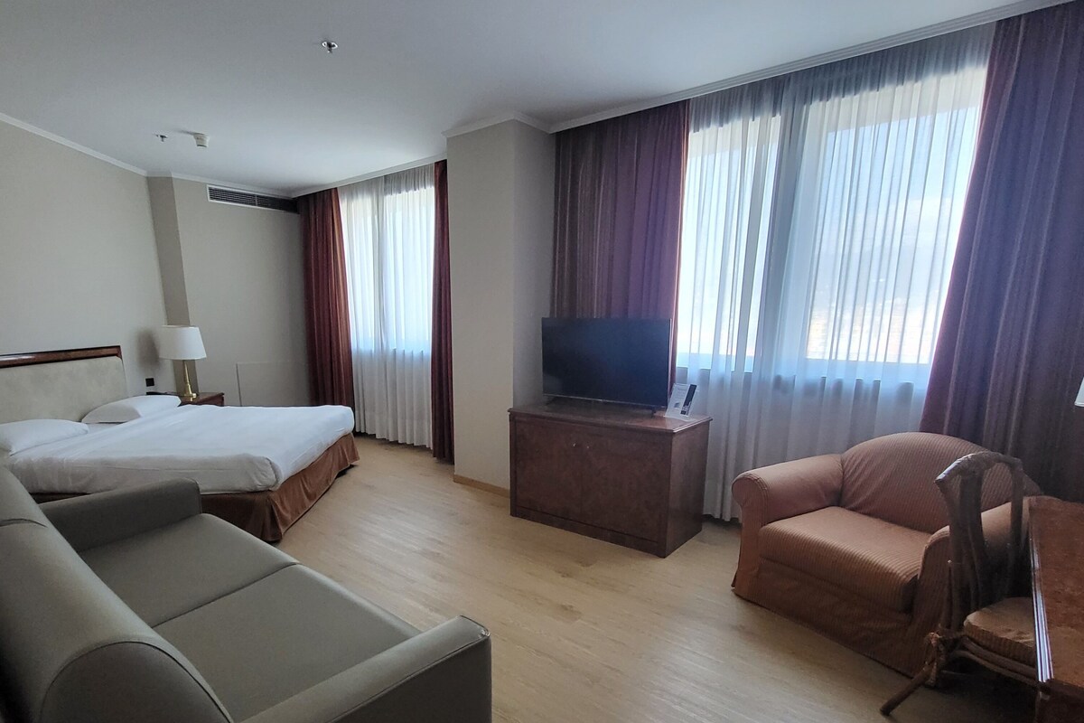A hotel room with a bed, a sofa, a TV on a stand, and a window with curtains.