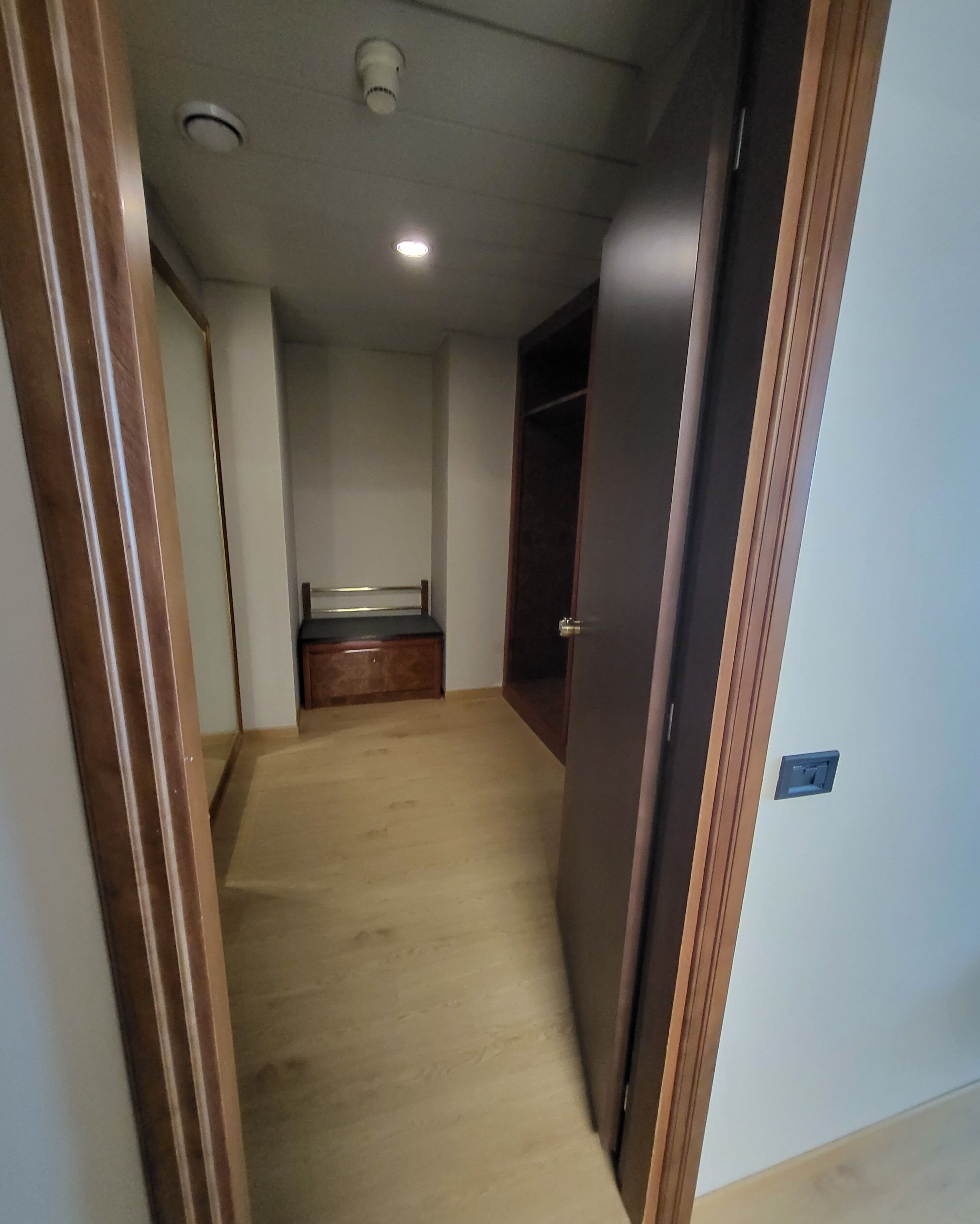 A narrow hallway with a wooden floor, a bed in the distance, and a door on the right.