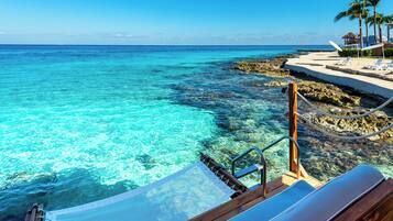 Vista despejada de una playa con muelle de madera y sillones de descanso, con vista a aguas turquesas y una costa rocosa.