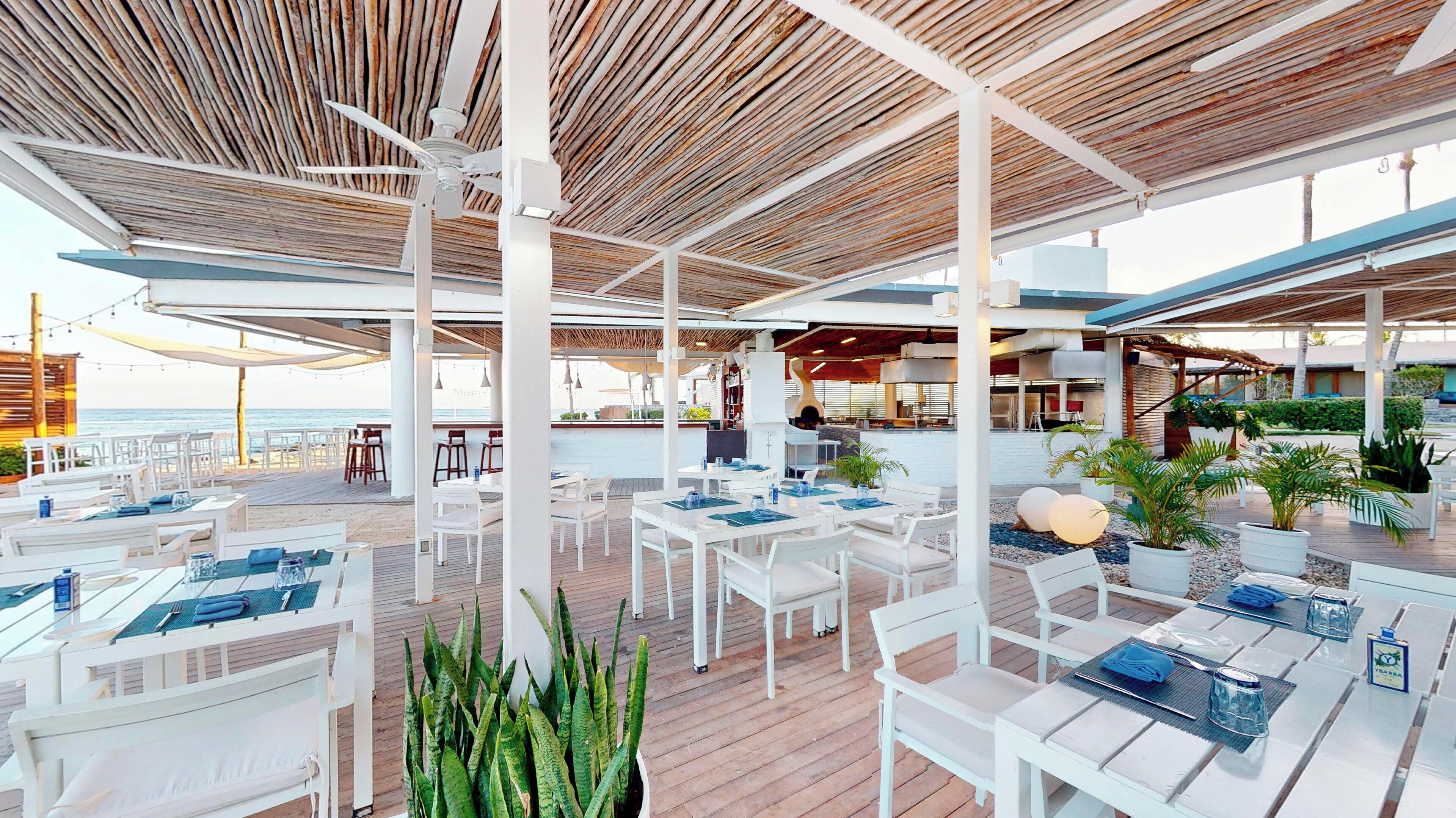 Un espace de restauration en plein air avec des tables et des chaises blanches, un plafond en bois et une vue sur l’océan.