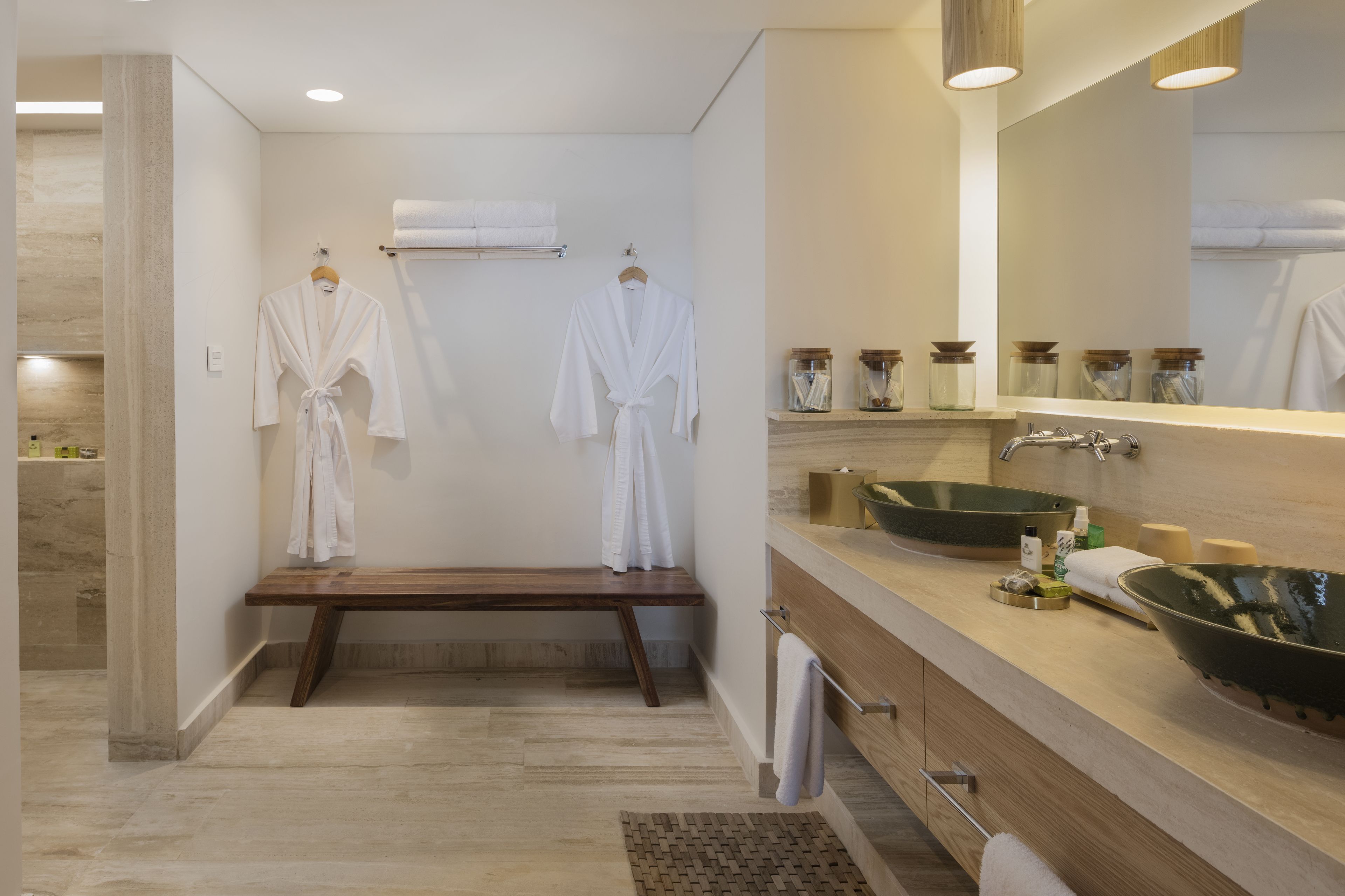 Un baño moderno con un banco de madera, dos albornoces blancos colgados en ganchos, un espejo grande y un lavabo con lavabo tipo cuenco.