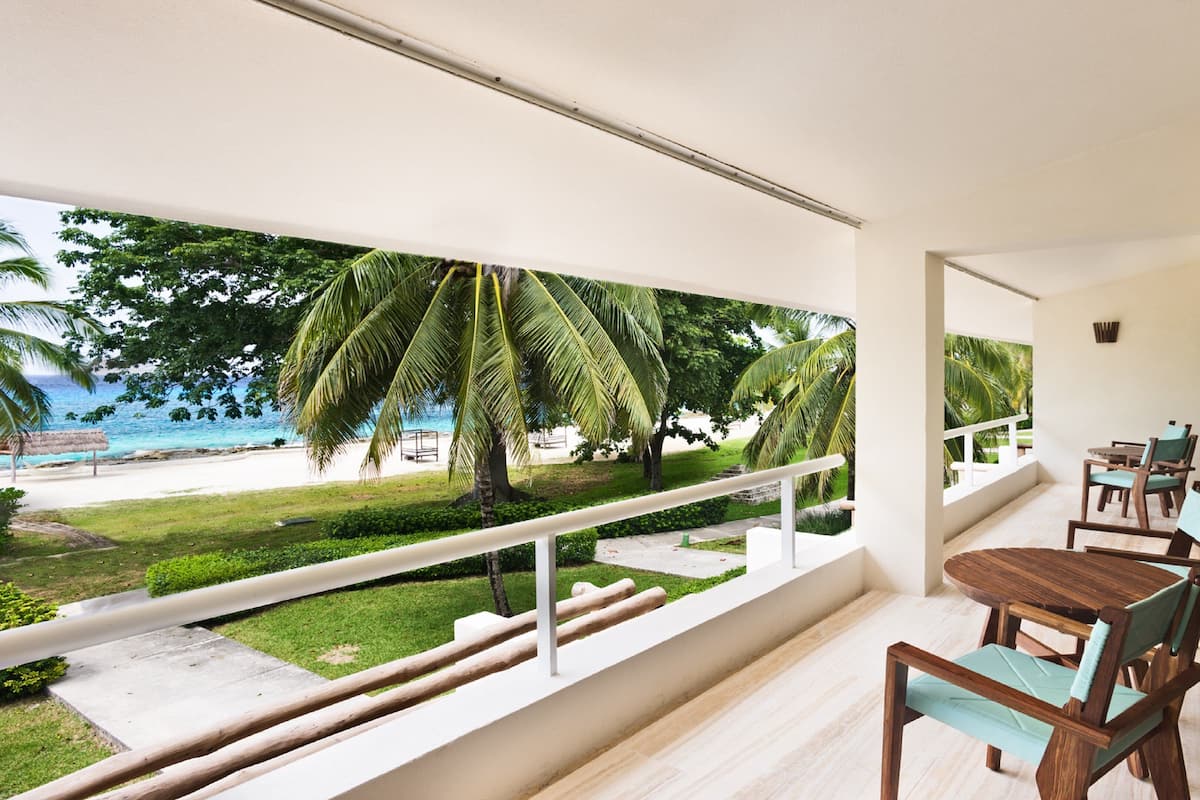 Un balcon avec une table et des chaises en bois, donnant sur une plage bordée de palmiers et offrant une vue dégagée sur l’océan.