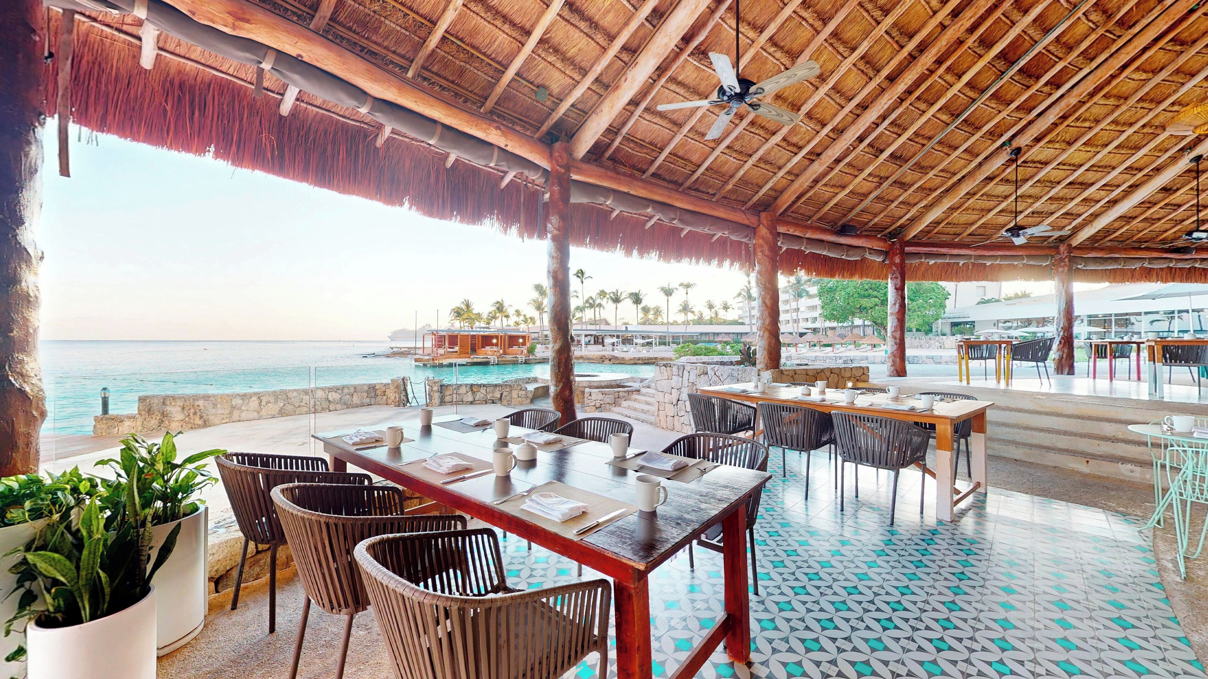 An outdoor dining area with a view of the sea, wooden tables, chairs, and a thatched roof.