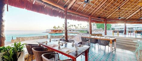 An outdoor dining area with a view of the sea, wooden tables, chairs, and a thatched roof.