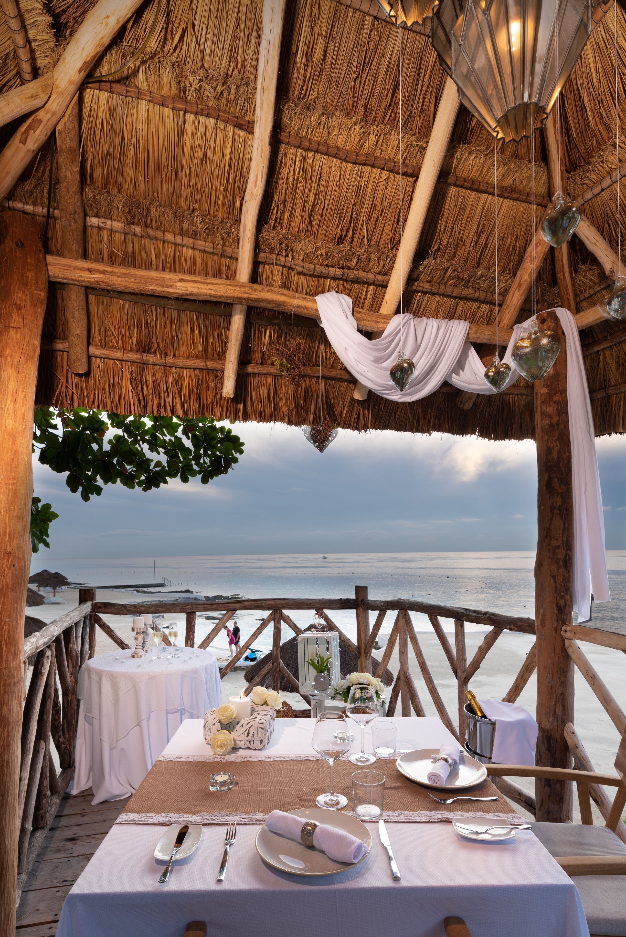 A dining table set for two with a view of the ocean, a thatched roof, and a wooden deck.