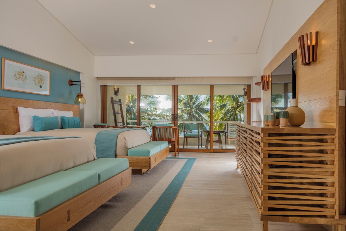 A modern bedroom with a large bed, a sofa, a desk, and a balcony with a view of palm trees.