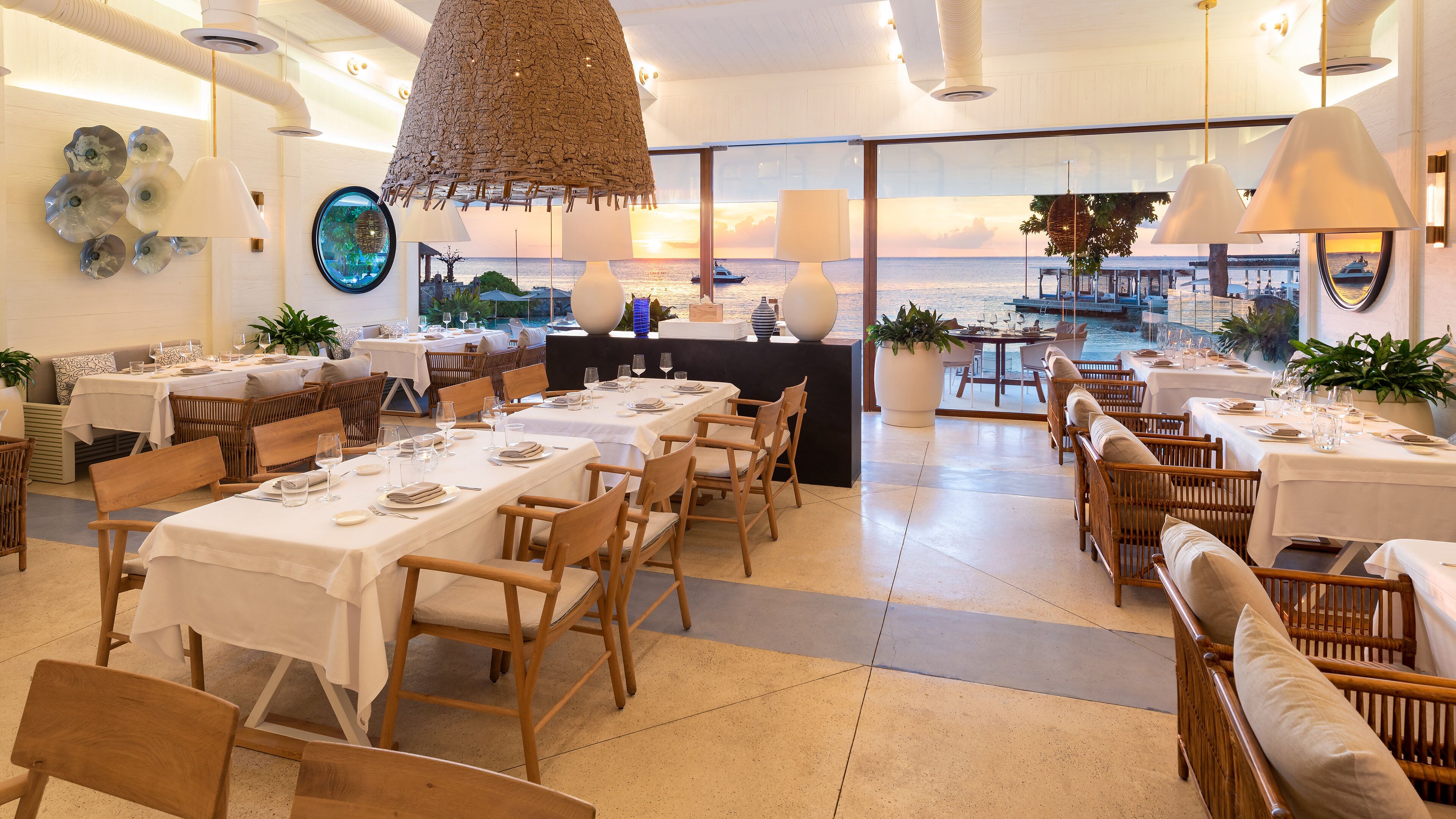 A restaurant with tables set for dining, a view of the sea, and a hanging light fixture.
